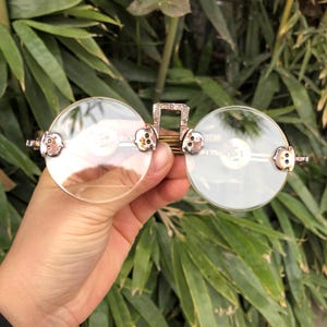 May include: Round eyeglasses with clear lenses and a gold-colored metal frame. The frame features decorative accents on the temples and bridge. The glasses are held against a backdrop of green foliage.