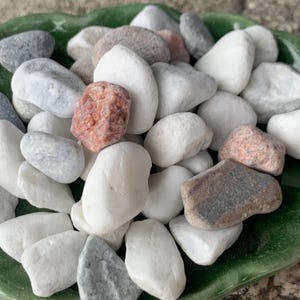 May include: A selection of smooth, polished stones in shades of white, grey, and reddish-brown. The stones vary in size and shape, and are presented in a green, leaf-shaped dish. Suitable for decorative use, these stones offer a natural aesthetic.