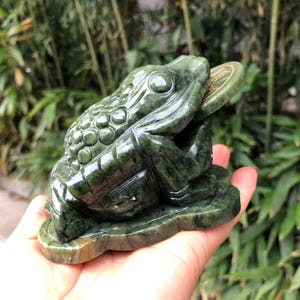 May include: A dark green jade frog sculpture holding a coin in its mouth. The frog is detailed with textured skin and sits on a base. The coin has gold-colored accents. The sculpture is held in a hand.