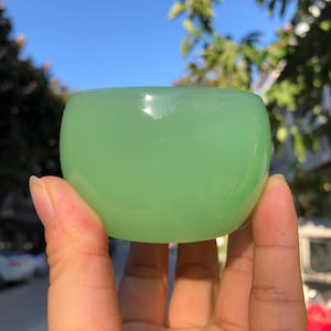 May include: A light green, rounded bowl with a smooth surface. The bowl is held in a hand, with a clear blue sky and green foliage in the background. The bowl has a glossy finish.