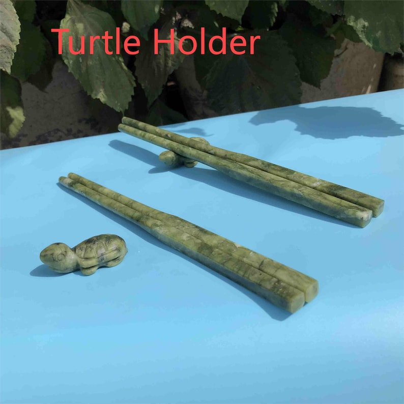 Op de afbeelding: Groene stenen eetstokjes en een schildpadvormige eetstokjeshouder op een blauw oppervlak. De eetstokjes zijn rechthoekig met een textuur. De tekst "Turtle Holder" staat in het rood bovenaan.