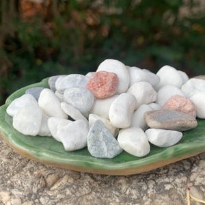 May include: A selection of smooth, polished stones in shades of white, grey, and reddish-brown, presented on a small, green, leaf-shaped ceramic dish. The stones are of varying sizes and shapes, offering a natural, textured aesthetic.