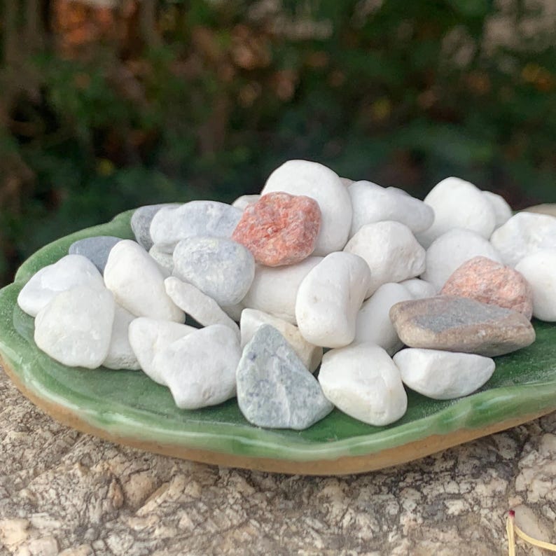 May include: A selection of smooth, polished stones in shades of white, grey, and reddish-brown, presented on a small, green, leaf-shaped ceramic dish. The stones are of varying sizes and shapes, offering a natural, textured aesthetic.