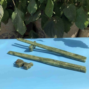 May include: Two pairs of green stone chopsticks with matching chopstick rests. The chopsticks are a light green color with darker green marbling. The chopstick rests are small, sculpted figures. The background is a light blue surface with green foliage.
