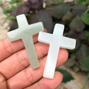 May include: Two jade crosses, one pale green and one white, are held in a hand. The crosses have a simple, classic design, with a smooth, polished surface. The background is blurred, with green foliage.