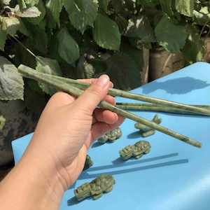 Op de afbeelding: Groene jade eetstokjes worden door een hand vastgehouden, met andere jade sculpturen van schildpadden en wezens op een blauw oppervlak. De eetstokjes zijn ongeveer 23 cm lang.