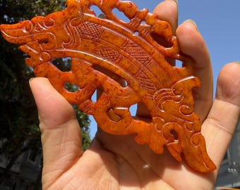 Op de afbeelding: Een gesneden oranjerode jade hanger met ingewikkeld opengewerkt en gedetailleerde patronen. De hanger wordt in een hand gehouden tegen een achtergrond van een blauwe lucht en boomtakken. Het ontwerp heeft een gestileerde, vloeiende vorm.