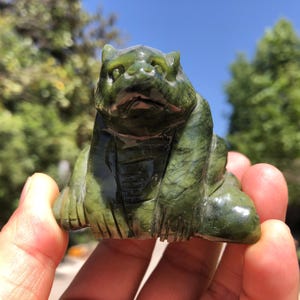 May include: A carved green jade figurine of a seated bear with detailed features. The bear has a slightly furrowed brow and is posed with its paws resting in front. The jade has natural variations in color.