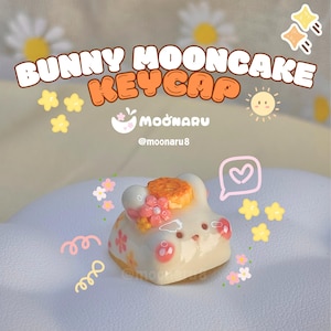 May include: A white and pink Bunny Mooncake keycap with a floral design. The keycap has a bunny face with a mooncake on its head. The text "BUNNY MOONCAKE KEYCAP" is displayed above the keycap.