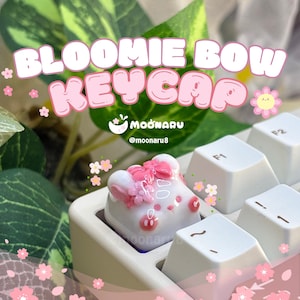 May include: A white and pink Bloomie Bow keycap with a bow and flower design, installed on a white keyboard. The keycap has a cartoon-like aesthetic. The text "Bloomie Bow Keycap" is displayed above the keycap.