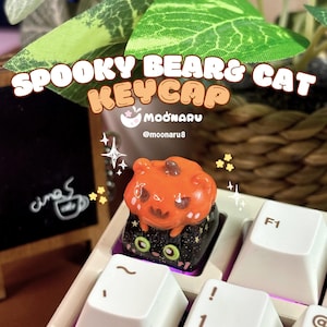 May include: A custom keycap featuring a spooky bear and cat design. The keycap is orange with a black base and is placed on a white keyboard. The text "SPOOKY BEAR & CAT KEYCAP" is displayed above the keycap.