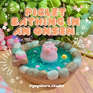Puede incluir: Diorama en miniatura con un cerdito rosa relajándose en un onsen turquesa, rodeado de piedras. La escena incluye un pequeño pato amarillo, accesorios de spa en miniatura y el texto "PIGLET BATHING IN AN ONSEN". El nombre del artista también es visible.