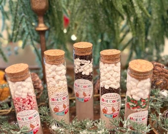 May include: Five glass test tubes filled with layered hot cocoa mix ingredients, each topped with a cork stopper. The tubes contain white marshmallows, sprinkles, and cocoa powder, with labels reading Hot Cocoa and instructions for preparation.