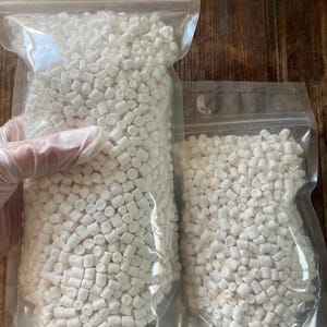 Puede incluir: Dos bolsas de plástico transparente llenas de pequeños objetos cilíndricos blancos. Las bolsas están selladas y parecen contener un producto alimenticio, posiblemente malvaviscos o una confitería similar. Las bolsas están sobre una superficie de madera.