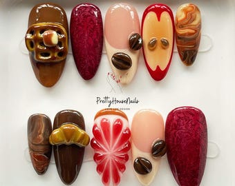 May include: A set of ten artificial nails with various designs. The nails feature coffee bean, fruit, and patterned designs in shades of brown, red, and peach. The text "Pretty House Nails" is visible.
