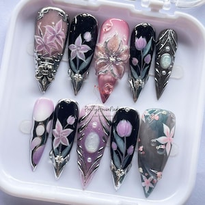 May include: A set of ten long, pointed artificial nails with intricate floral designs. The nails feature a variety of colors, including pink, black, and silver, with detailed flower and leaf patterns. The text "PrettyHouseNails" is visible.