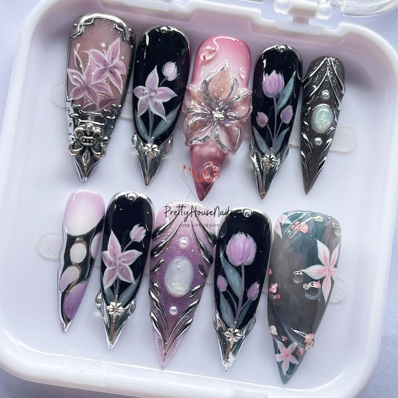 May include: A set of ten long, pointed artificial nails with intricate floral designs. The nails feature a variety of colors, including pink, black, and silver, with detailed flower and leaf patterns. The text "PrettyHouseNails" is visible.