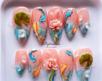May include: A set of ten artificial nails with a pink base, featuring 3D designs of koi carp, lotus flowers, and lily pads. The nails have blue and white water effects. The text "Pretty House Nails" is visible.