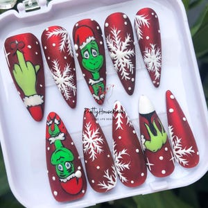 May include: Set of ten red and white Christmas-themed false nails with a Grinch design. The nails feature snowflakes, white dots, and a green Grinch character.