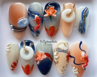 May include: A set of ten almond-shaped fake nails with various designs. The designs include a white and blue floral pattern, a red and gold floral pattern, a red and gold fish pattern, and a white and gold cloud pattern. The nails are arranged in a row on a white surface.