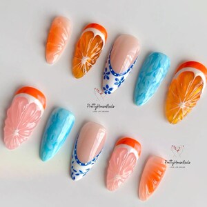 May include: Set of ten false nails with various designs. Some nails feature a 3D orange slice design, while others have a blue and white floral design. The nails are all in a stiletto shape.