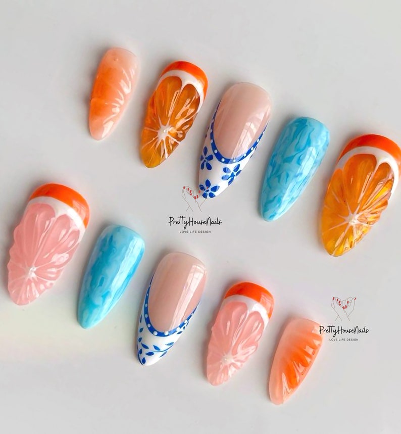 May include: Set of ten false nails with various designs. Some nails feature a 3D orange slice design, while others have a blue and white floral design. The nails are all in a stiletto shape.