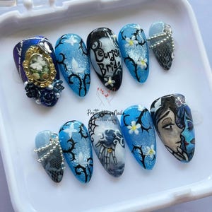 May include: A set of ten long, oval-shaped artificial nails. The nails feature various designs, including floral patterns, spiderwebs, and the text "Corpse Bride." Colors include blue, black, white, and silver. Some nails have 3D embellishments.