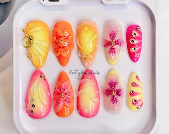 Puede incluir: Conjunto de diez uñas postizas con un diseño degradado en rosa, amarillo y naranja. Cada uña presenta una flor 3D con pedrería y detalles dorados. Las uñas se muestran en una caja blanca con el texto "PrettyHouseNails LOVE LIFE DESIGN."