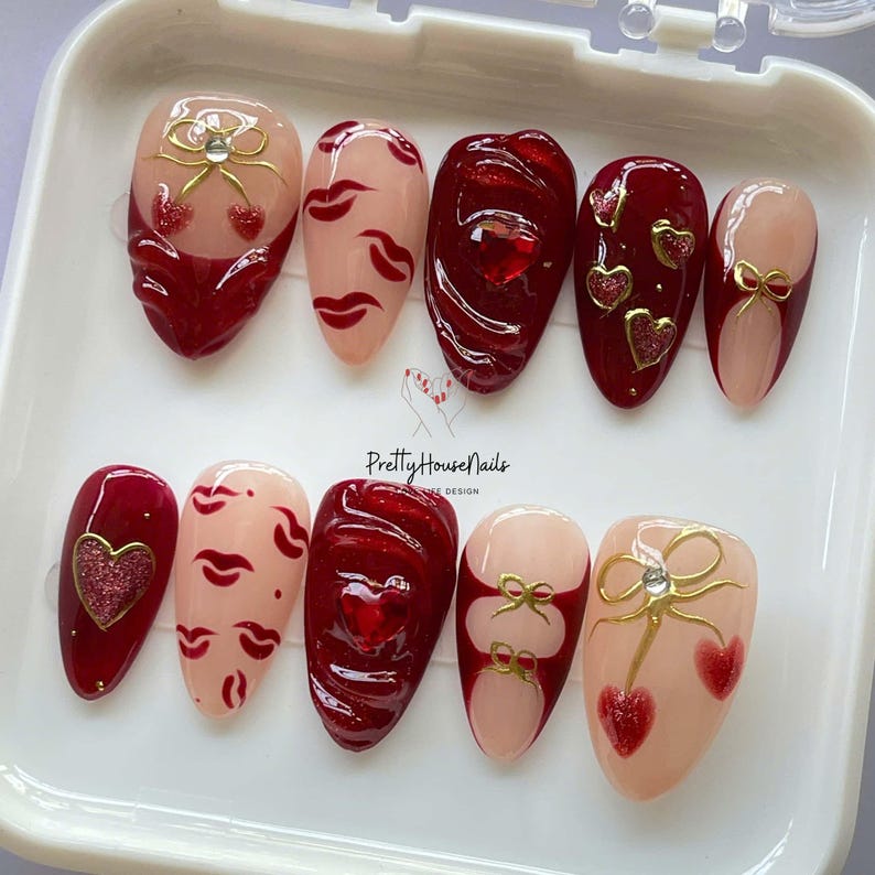 May include: A set of ten artificial nails with various designs. The nails feature a combination of red, nude, and gold accents. Designs include hearts, bows, cherries, and lip prints. The text "Pretty House Nails" is visible.