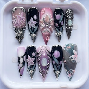 May include: A set of ten long, pointed false nails. Each nail features intricate designs, including floral patterns, gems, and metallic accents. Colors include pink, black, purple, and silver, creating a glamorous and artistic aesthetic.
