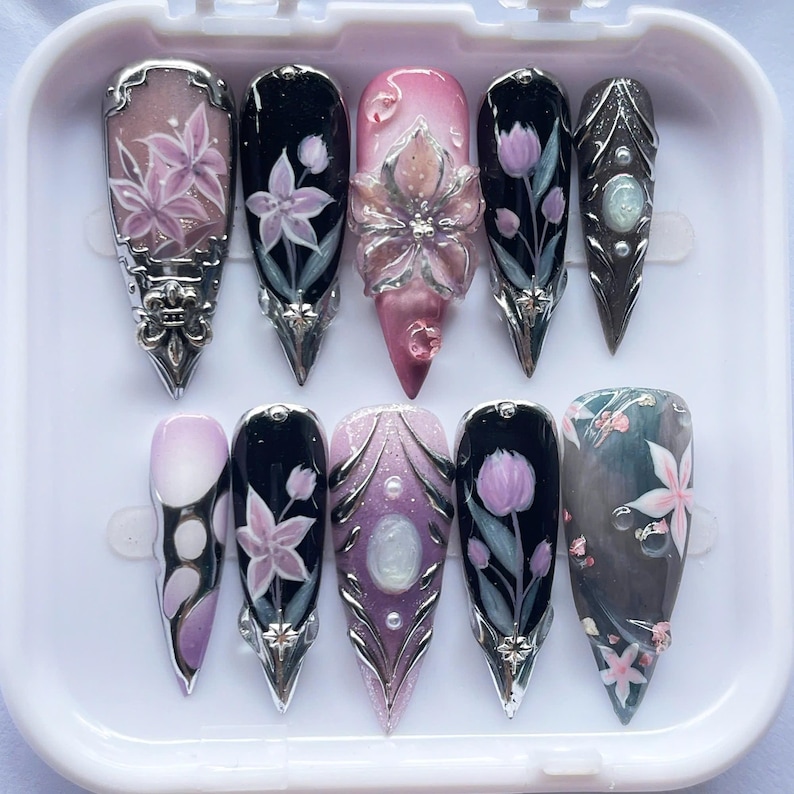 May include: A set of ten long, pointed false nails. Each nail features intricate designs, including floral patterns, gems, and metallic accents. Colors include pink, black, purple, and silver, creating a glamorous and artistic aesthetic.
