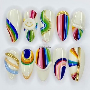 Pode incluir: Dez unhas postiças brancas ovais com diferentes desenhos de arco-íris. Algumas unhas têm um rosto sorridente, um redemoinho e uma faixa dourada.