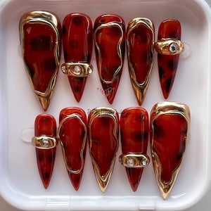 Può includere: Set di dieci unghie finte con un design a scaglie di tartaruga nei toni del rosso e del marrone. Ogni unghia è rifinita con bordi dorati e un anello dorato con una piccola gemma trasparente. Presentate in un contenitore bianco.