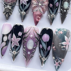 May include: A collection of elaborately designed artificial nails. Each nail features a unique design, including floral patterns, glitter, and metallic accents. The color palette includes shades of pink, purple, black, and silver, creating a glamorous aesthetic.
