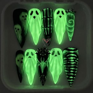 May include: A set of ten long, pointed artificial nails with a Halloween theme. Designs include glowing green ghosts, a skeleton, and swirling patterns. The nails are in a clear, rectangular container.