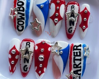 Puede incluir: Un conjunto de diez uñas artificiales con varios diseños. Las uñas presentan combinaciones de colores rojo, blanco y azul, adornos en forma de estrella y textos que dicen "COWBOY", "BANG" y "CARTER".