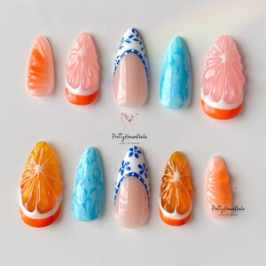 May include: Set of ten false nails with various designs. Some nails feature a 3D orange slice design with a white outline and orange base. Other nails have a blue and white floral design with a white base. The nails are arranged on a white surface.