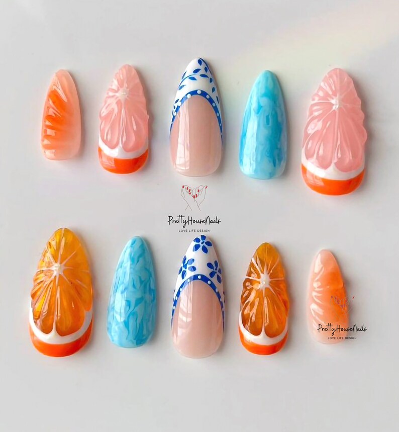 May include: Set of ten false nails with various designs. Some nails feature a 3D orange slice design with a white outline and orange base. Other nails have a blue and white floral design with a white base. The nails are arranged on a white surface.
