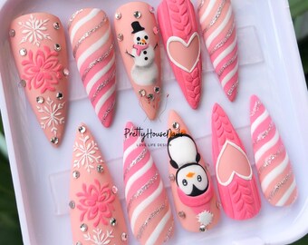 Pode incluir: Conjunto de dez unhas postiças rosa e brancas com designs de tema de inverno. Os designs incluem um boneco de neve, um pinguim, flocos de neve e um coração. As unhas estão dispostas em uma caixa branca.