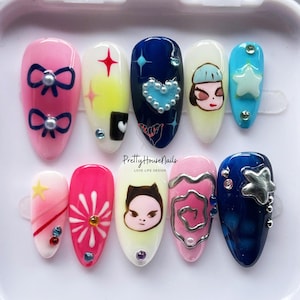 May include: Set of ten false nails with various designs. The nails are painted with different colors and patterns, including pink, blue, yellow, and white. Some nails feature stars, hearts, flowers, and cartoon characters. The nails are arranged in two rows on a white surface.