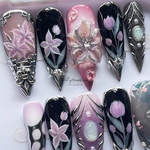 May include: A collection of long, pointed artificial nails with intricate floral designs. The nails feature a variety of colors, including pink, black, and silver, with detailed artwork of flowers and embellishments. The text "Pretty House Nails LOVE LIFE DESIGN" is visible.