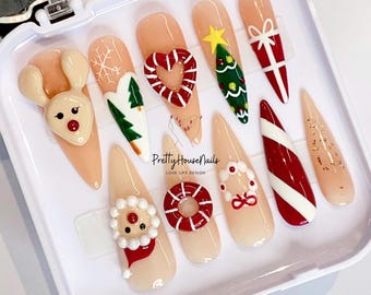 Pode incluir: Um conjunto de dez unhas postiças com tema natalino com vários designs, incluindo uma rena, uma árvore de Natal, um bastão de caramelo, um homem de gengibre e uma coroa. As unhas são todas de cor rosa claro e estão dispostas em uma caixa branca.