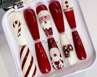 Pode incluir: Um conjunto de dez unhas postiças de Natal vermelhas e brancas em uma caixa branca. As unhas apresentam desenhos de bengalas de doces, Papai Noel, renas e flocos de neve.