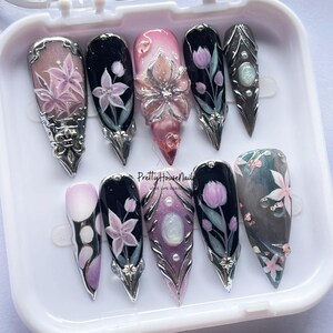 May include: A set of ten artificial nails with various designs. The nails feature floral patterns, glitter, and metallic accents. Some nails have a 3D flower design. The color palette includes pink, black, silver, and purple. The text "Pretty House Nails" is visible.
