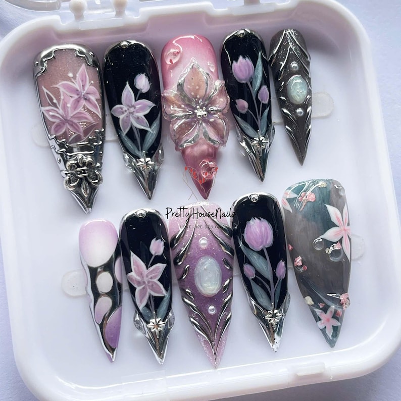 May include: A set of ten artificial nails with various designs. The nails feature floral patterns, glitter, and metallic accents. Some nails have a 3D flower design. The color palette includes pink, black, silver, and purple. The text "Pretty House Nails" is visible.