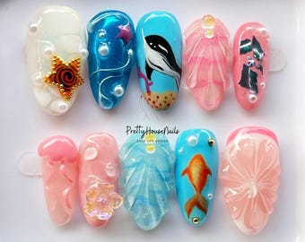 Puede incluir: Un conjunto de diez uñas postizas con un diseño de temática marina. Las uñas están pintadas en tonos de azul, rosa y blanco, y presentan varias criaturas marinas, incluyendo una ballena, una medusa, una estrella de mar y una concha. Las uñas están dispuestas sobre una superficie blanca.