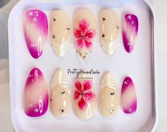 May include: A set of ten false nails in a white box. The nails are oval shaped and have a gradient design with pink and white. Some of the nails have a 3D flower design with pink and white accents. The box has the text "PrettyHouseNails LOVE LIFE DESIGN" printed on it.