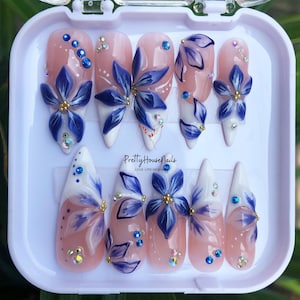 Puede incluir: Un conjunto de 10 uñas postizas con base blanca y rosa y diseños florales azules. Las uñas están dispuestas en un estuche de plástico blanco con el texto "PrettyHouseNails LOVE LIFE DESIGN" impreso en la parte inferior.