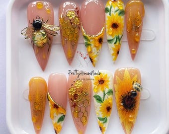 May include: A set of ten long, pointed artificial nails. The nails feature a variety of designs, including sunflowers, honeycombs, and a 3D bee. The color palette includes shades of yellow, orange, and pink, with gold accents. The text "Pretty House Nails" is visible.