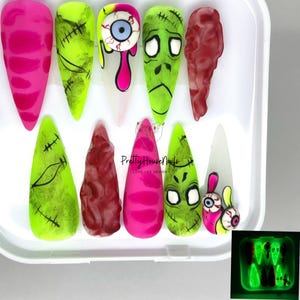 Puede incluir: Conjunto de 10 uñas postizas con temática de Halloween con varios diseños, incluyendo una cara de zombie verde, una uña a rayas rosa y blanca, una uña sangrienta roja y blanca y una uña verde con una boca cosida. Las uñas están en una caja de plástico blanca con una tapa transparente. Las uñas brillan en la oscuridad, como se muestra en la imagen de la derecha.